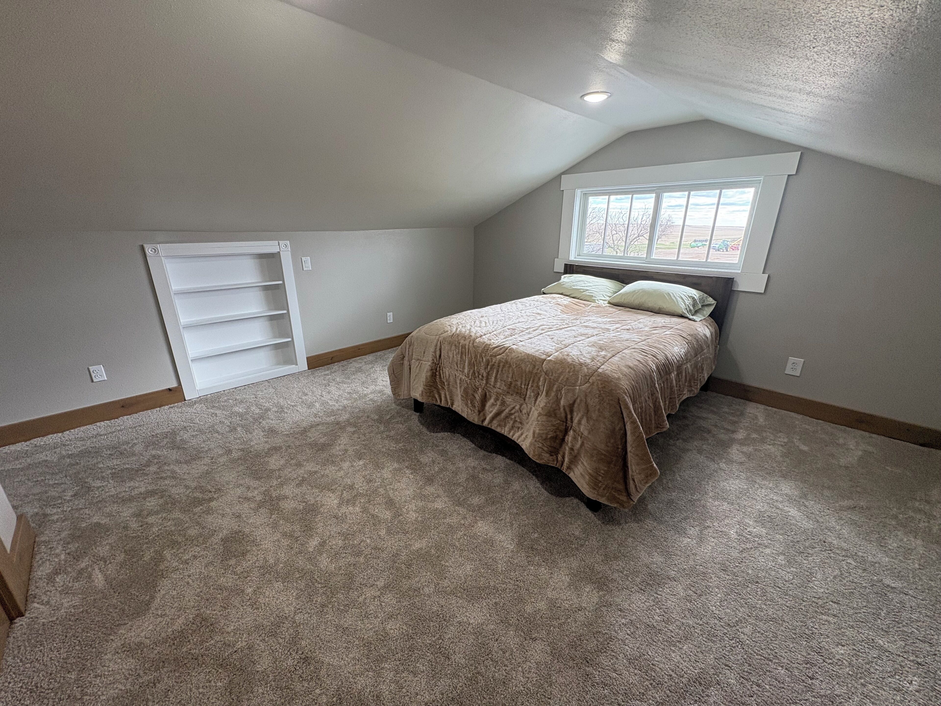 Une chambre avec un plafond en pente, un lit recouvert d’une couette beige, une fenêtre avec des stores et une étagère intégrée.