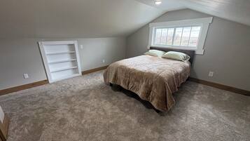 Une chambre avec un plafond en pente, un lit recouvert d’une couette beige, une fenêtre avec des stores et une étagère intégrée.
