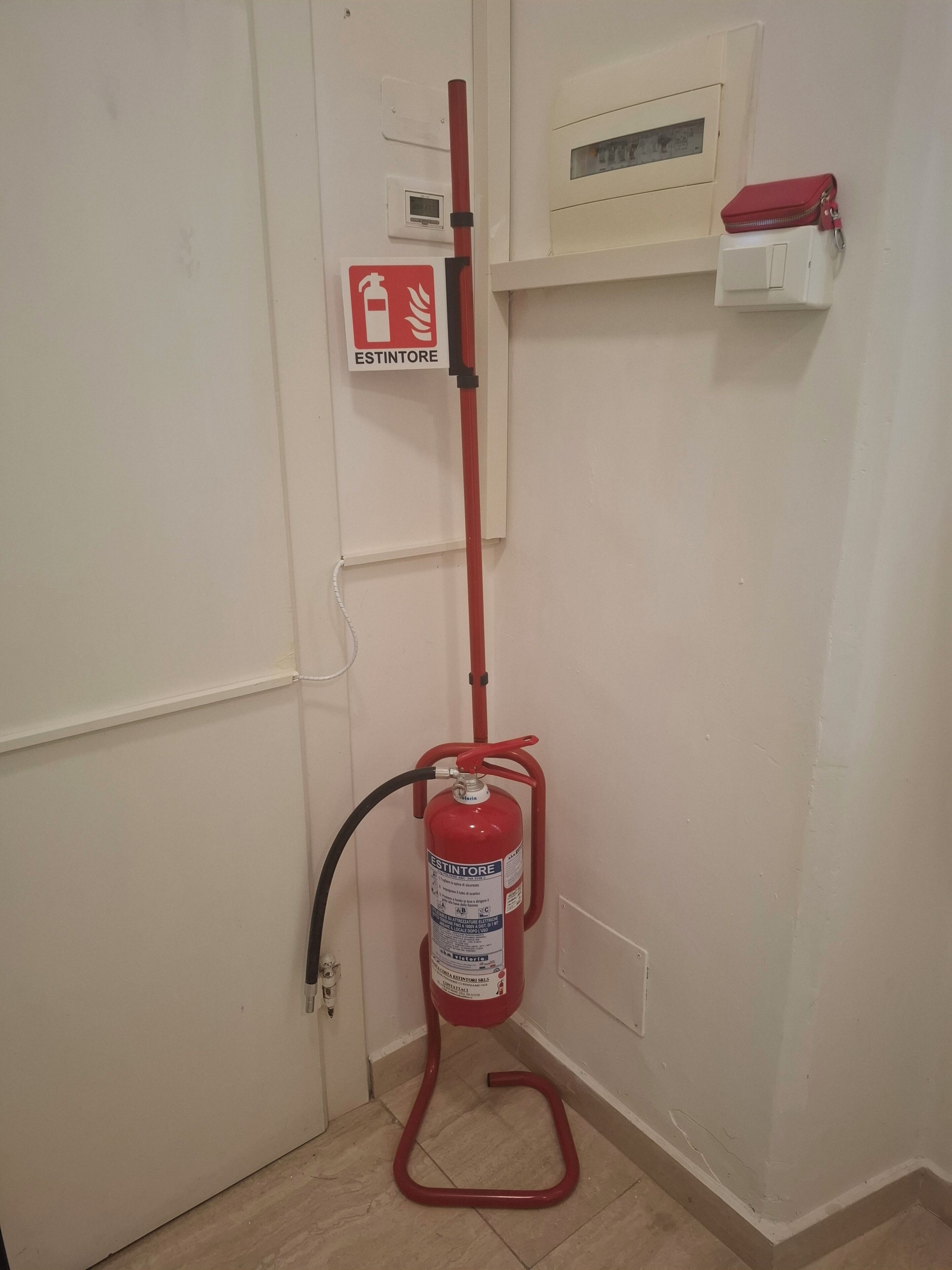 A fire extinguisher mounted on a wall, a fire alarm pull station, and a red bag on a shelf.