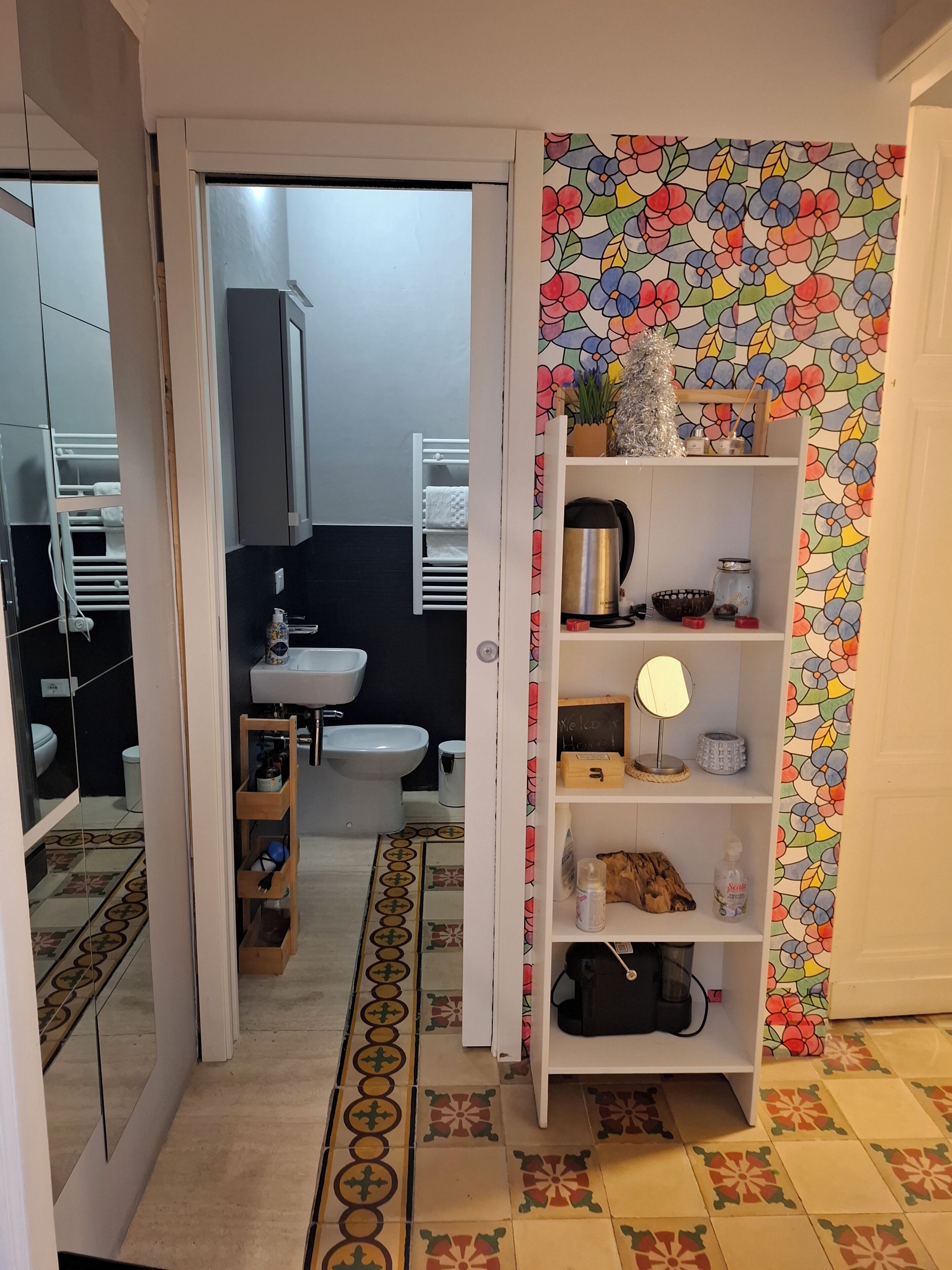 Un corridoio stretto con pavimento piastrellato, che conduce a un bagno con water, lavabo e bidet. Si nota uno scaffale bianco con diversi oggetti, e una carta da parati floreale colorata riveste la parete adiacente.