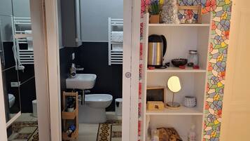 Un corridoio stretto con pavimento piastrellato, che conduce a un bagno con water, lavabo e bidet. Si nota uno scaffale bianco con diversi oggetti, e una carta da parati floreale colorata riveste la parete adiacente.