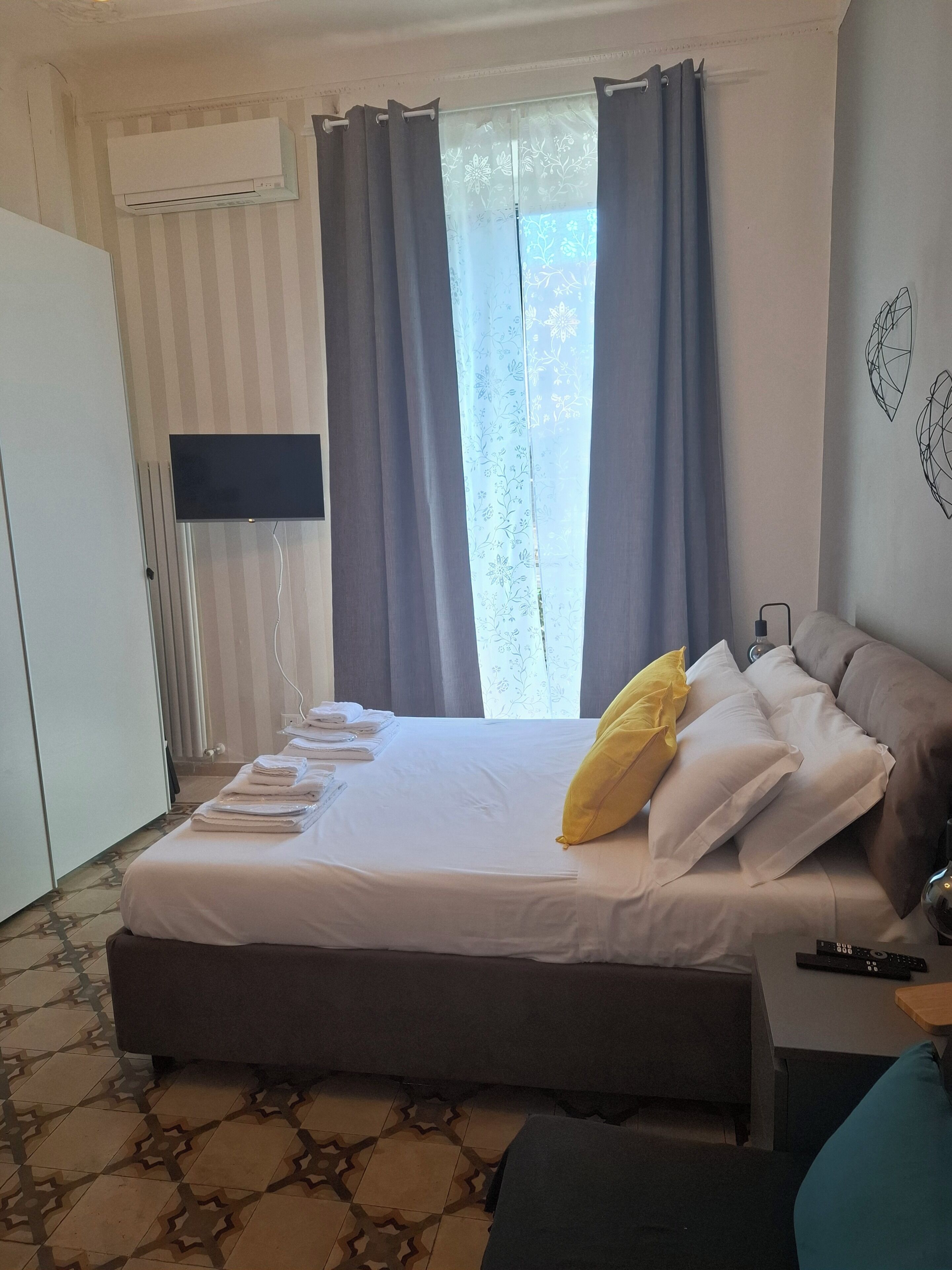 Habitación de hotel con cama, televisor de pantalla plana, mesita de noche, lámpara y ventana con cortinas.