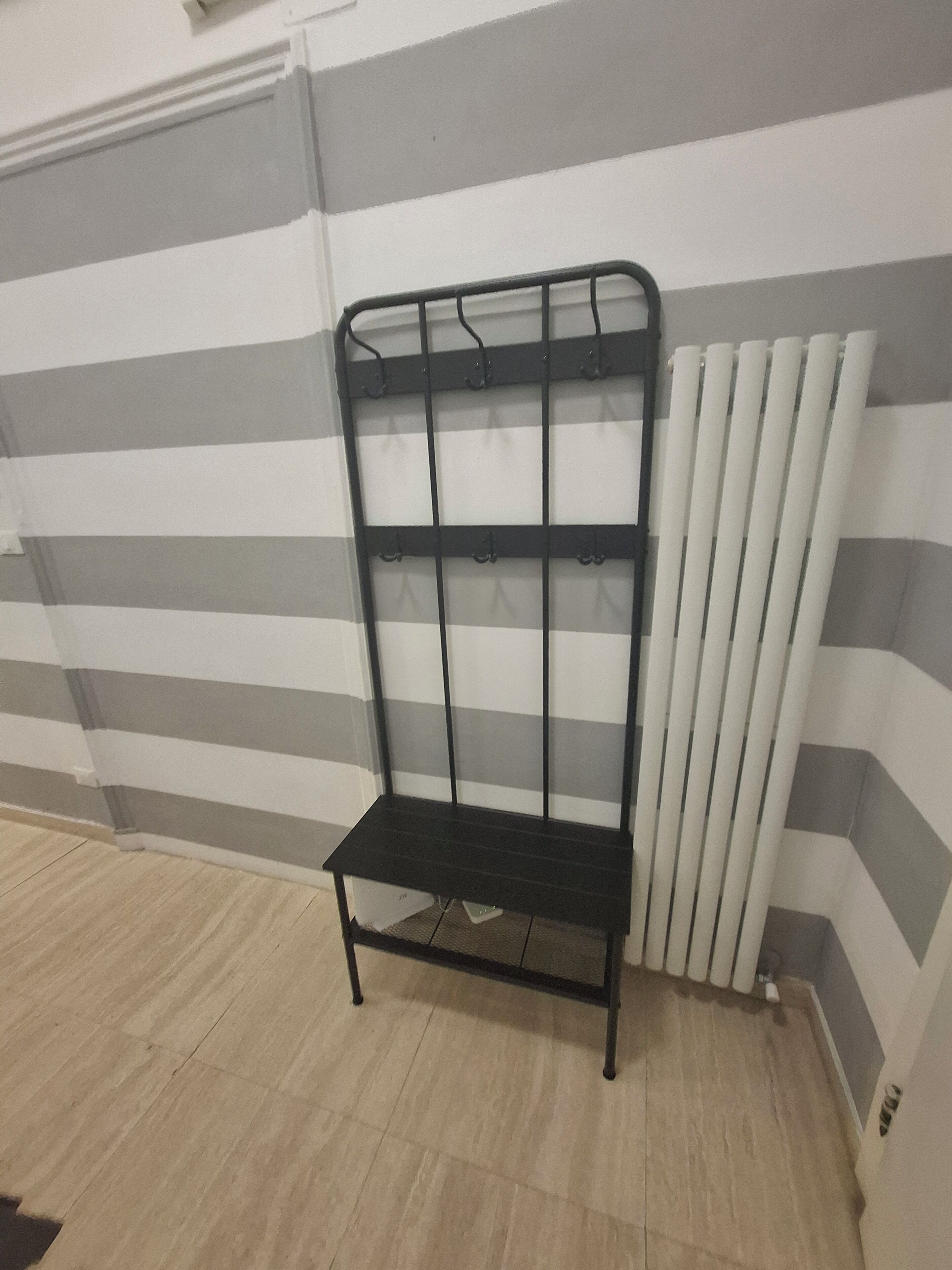 Ein schwarzes Metallregal mit drei Ablagen, vor einer Wand mit horizontalen Grau- und Weißstreifen platziert.