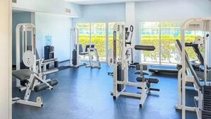 Un gimnasio con diferentes máquinas de ejercicio, piso azul y grandes ventanales con vistas a zonas verdes.