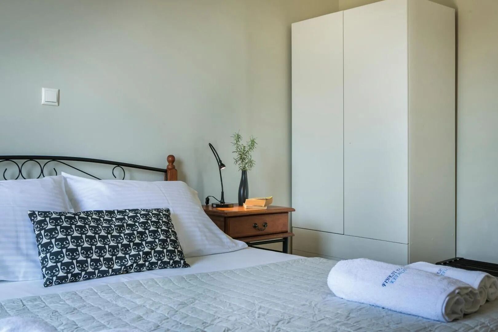 Una cama con ropa de cama blanca, una almohada estampada, una lámpara de noche, una planta pequeña y una toalla enrollada.