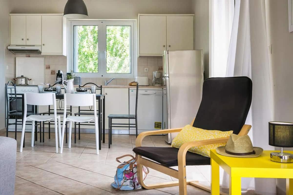 Una cocina compacta con armarios blancos, una zona de comedor con sillas blancas, una silla negra con cojín amarillo y una pequeña mesa amarilla con una lámpara.