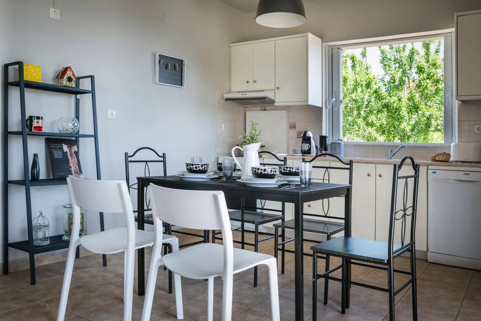A modern kitchen with white cabinets, a dining table with chairs, a window with a view of trees, and a shelf with decorative items.