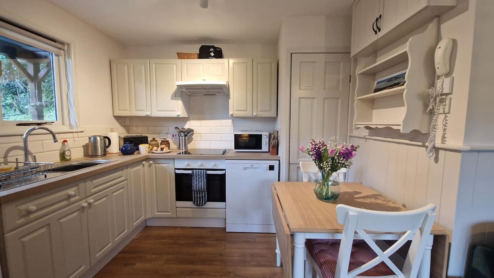 Une cuisine compacte avec des meubles blancs, un four à micro-ondes, un évier et une table à manger en bois accompagnée d’une chaise.