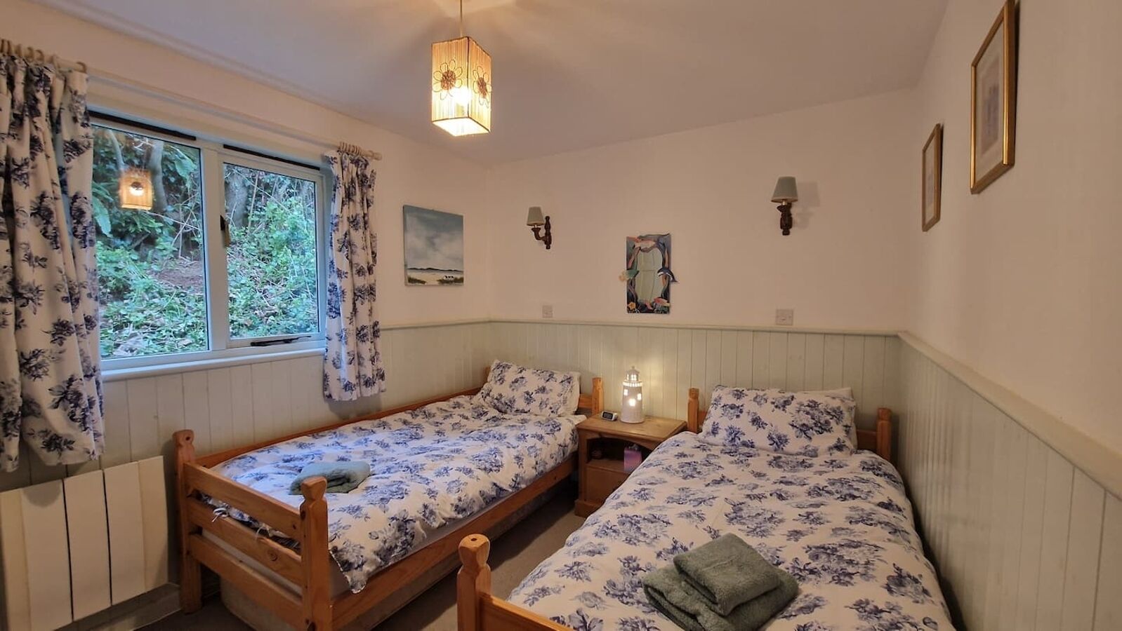 A room with two wooden beds, a window with curtains, a wall-mounted lamp, and a painting.