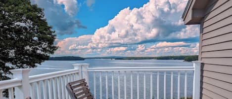 A wooden deck with white railings, overlooking a body of water with trees and a cloudy sky.