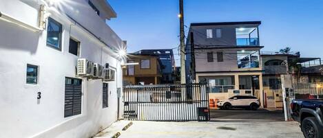 A gated parking area with a white building on the left and a two-story structure on the right.