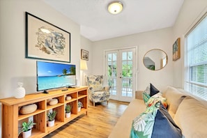 Ein Wohnzimmer mit Fernseher, einer Holz-Unterhaltungsecke, einem tropisch gestalteten Sessel und einem Sofa mit Blick ins Freie.