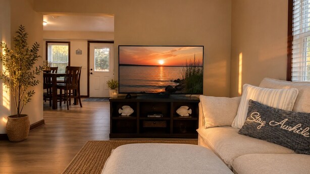 Un salon avec un téléviseur diffusant une image de coucher de soleil, un canapé avec un coussin, un coin repas avec des chaises et une plante en pot.