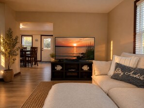 Una sala de estar con una televisión mostrando un atardecer, un sofá con un cojín, un comedor con sillas y una planta en maceta.