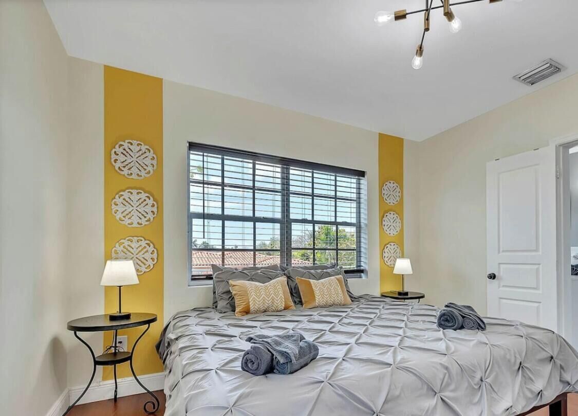 A bedroom with a large bed, two bedside tables with lamps, decorative wall art, a window with blinds, and a door.