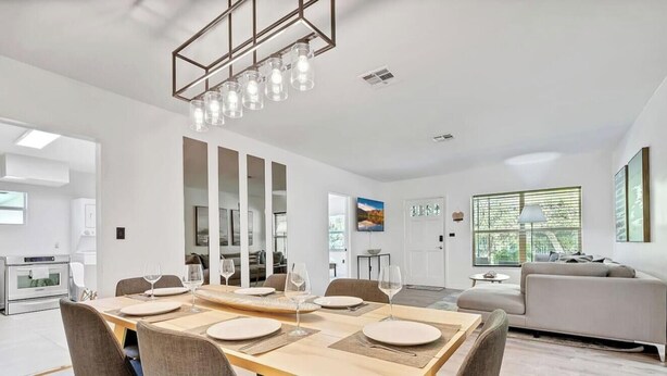 A modern dining area with a wooden table set for four, a kitchen in the background, and a living area with a sofa and a lamp. - 2-Home Compound with Pool and BBQ, Sleeps 16 (Miami)
