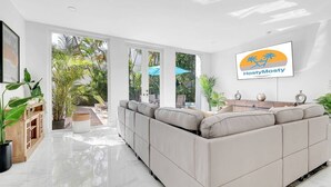 A modern living room with a large beige sofa, a TV mounted on the wall, and a view of a garden through large windows. - Gated Miami Luxury Oasis Pool & BBQ I 4-Bed, 4-Bath (Miami)