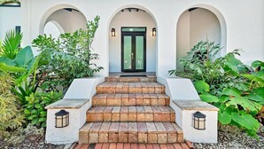 A white building with a black door, surrounded by greenery and featuring a set of stone steps leading to the entrance. - Gated Miami Luxury Oasis Pool & BBQ I 4-Bed, 4-Bath (Miami)