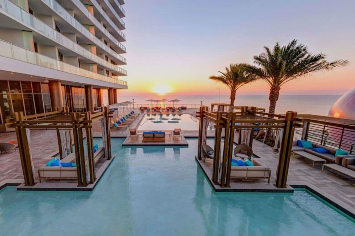 A resort pool area with sun loungers and a view of the sunset over the ocean.