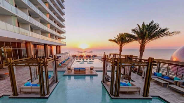 Zona de piscina en un resort con tumbonas y vista al atardecer sobre el océano.