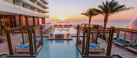 A resort pool area with sun loungers and a view of the sunset over the ocean.