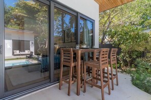 A patio with a table and chairs, a glass door leading to a pool, and a view of a house and trees. - Luxury Miami Oasis Heated Pool & BBQ with Guesthouse (Miami)