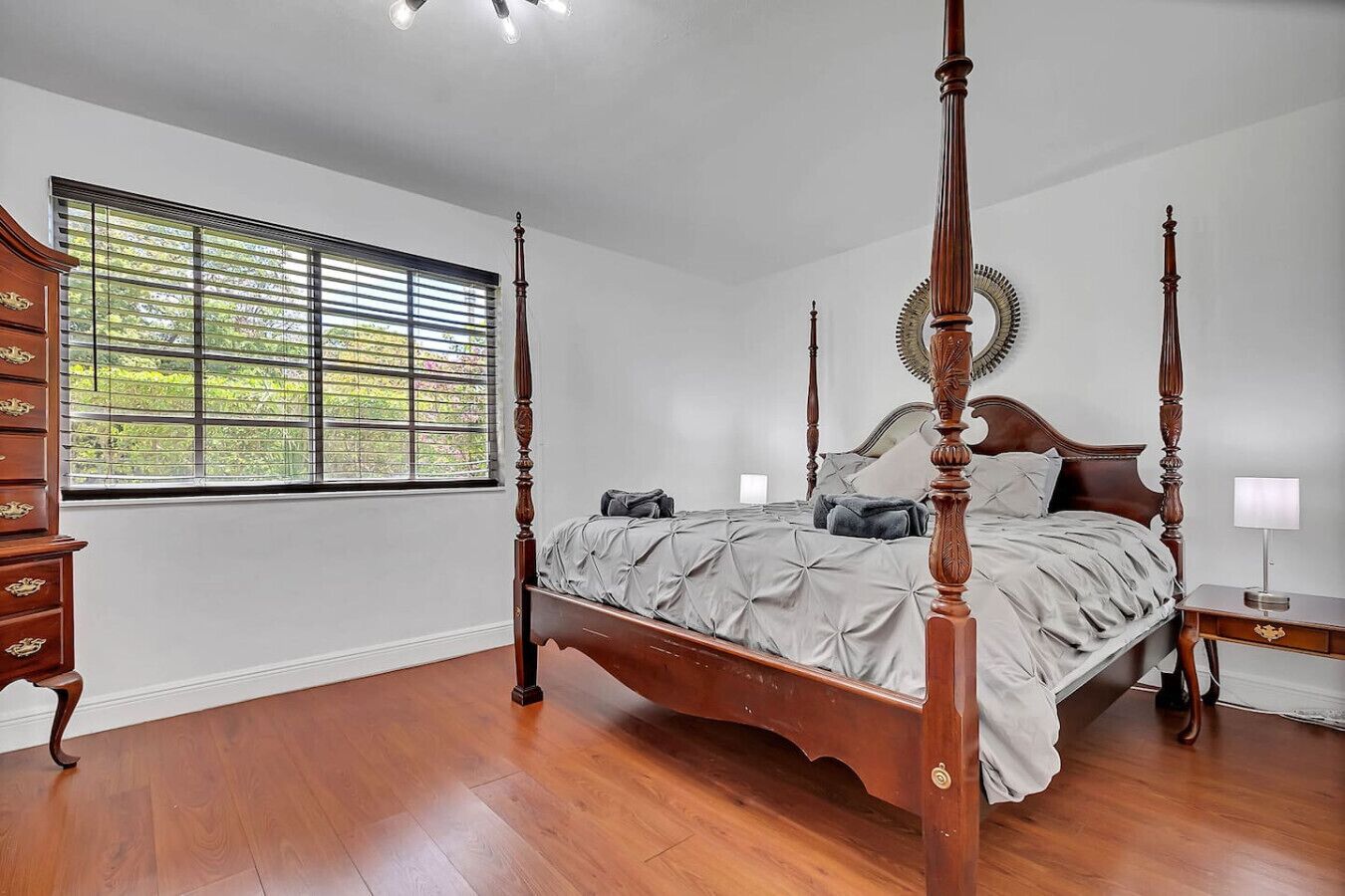 Ein Bett mit Baldachin, ein Holzbettgestell, eine Kommode mit Schubladen, ein Nachttisch mit Lampe und ein Fenster mit Jalousien.