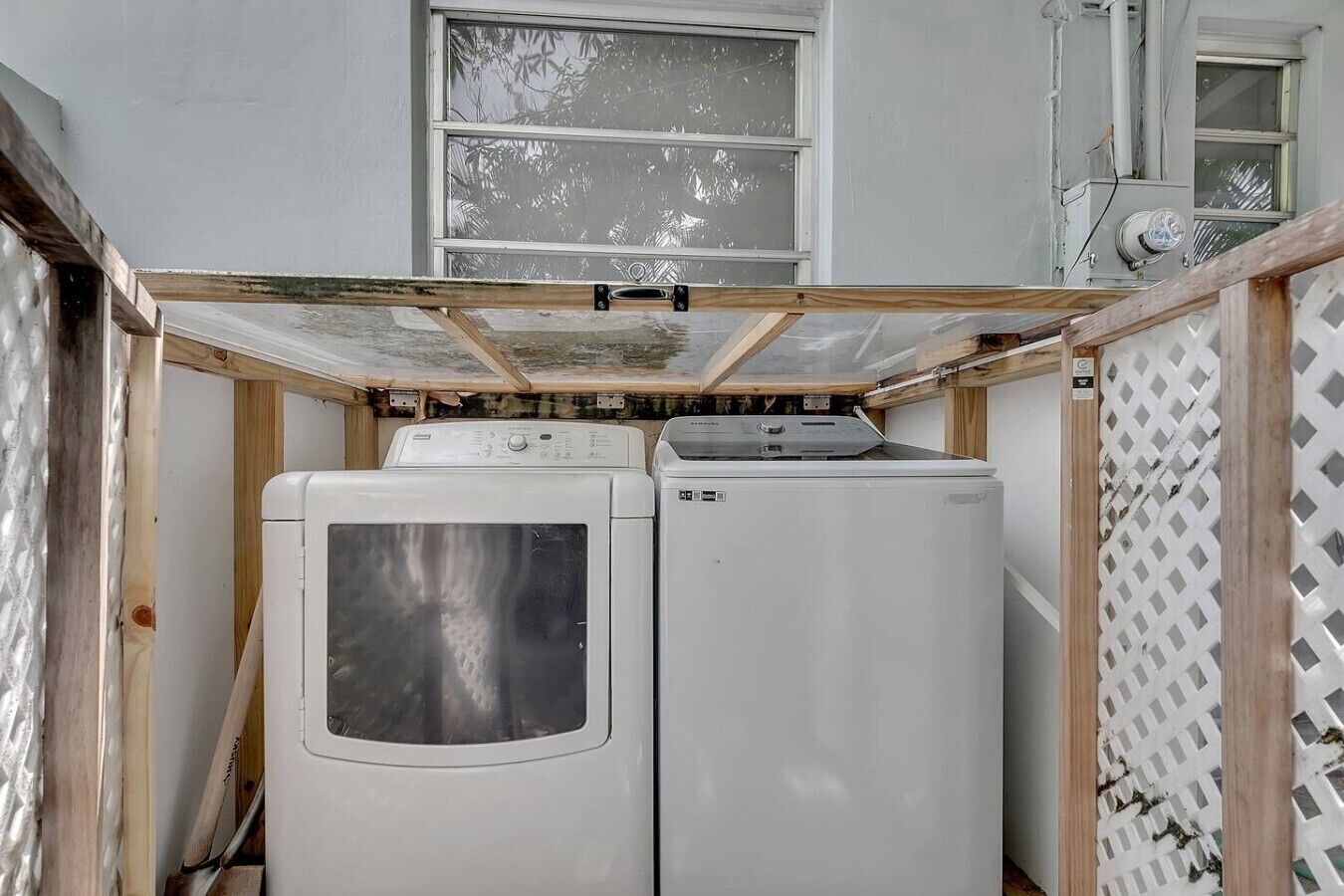 A laundry room with two white washing machines, a wooden ceiling, and a window with a view of trees.