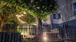 Un espace extérieur, éclairé la nuit, avec un arbre, une clôture en métal et un bâtiment doté d’une fenêtre.