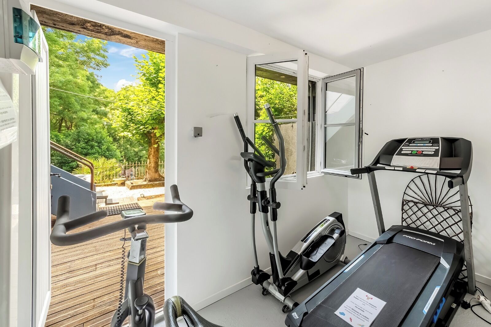 A home gym with an elliptical, a stationary bike, and a treadmill, with a view of a garden through a glass door.