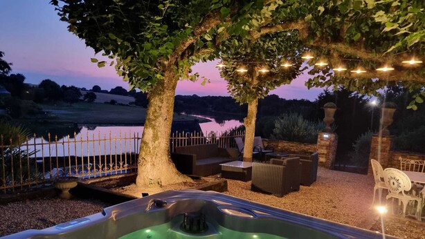 Un bain à remous avec vue sur un lac au crépuscule, entouré de meubles de jardin et illuminé de guirlandes lumineuses.
