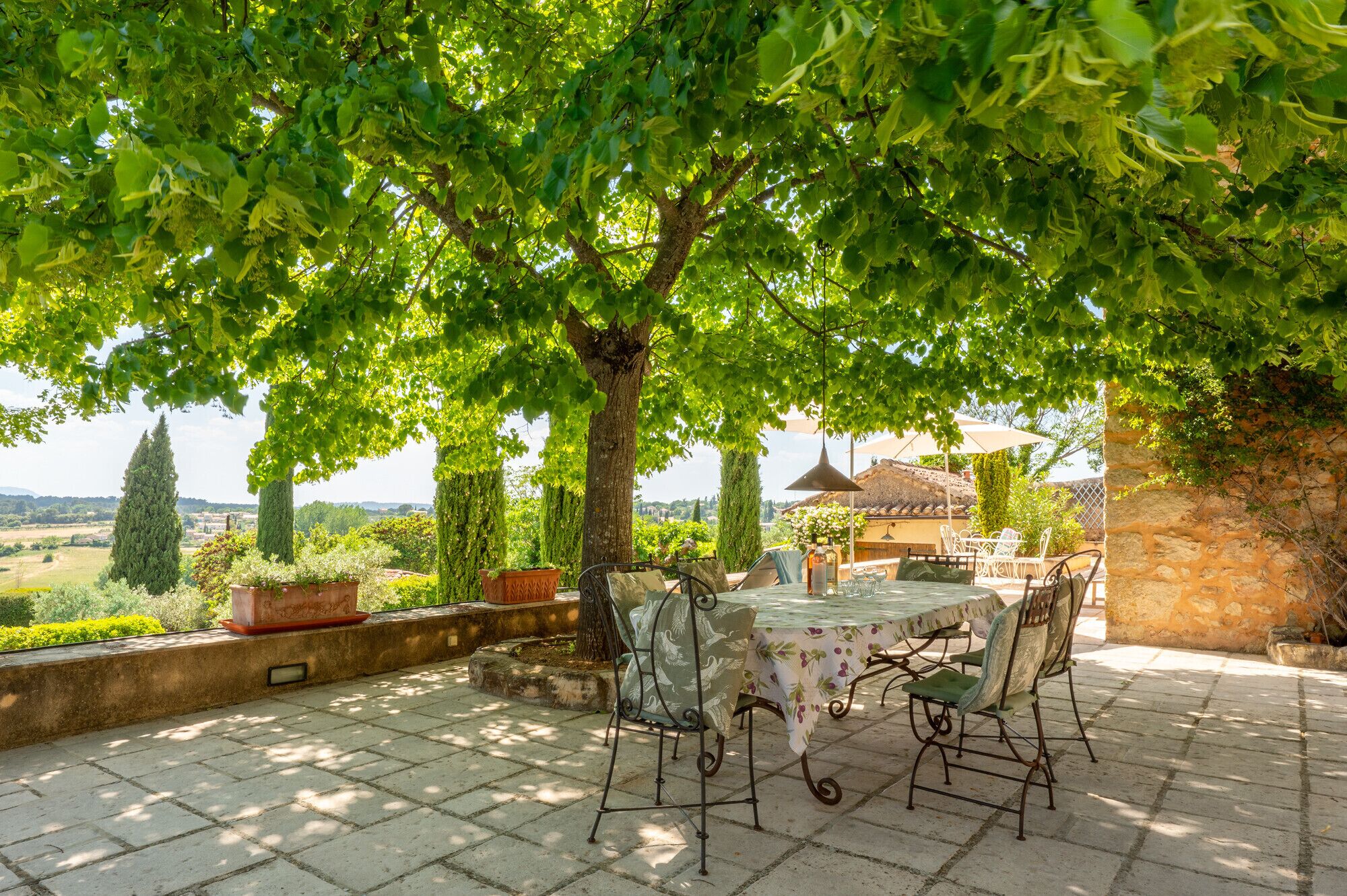 An outdoor patio with a table and chairs, surrounded by greenery and a stone wall.