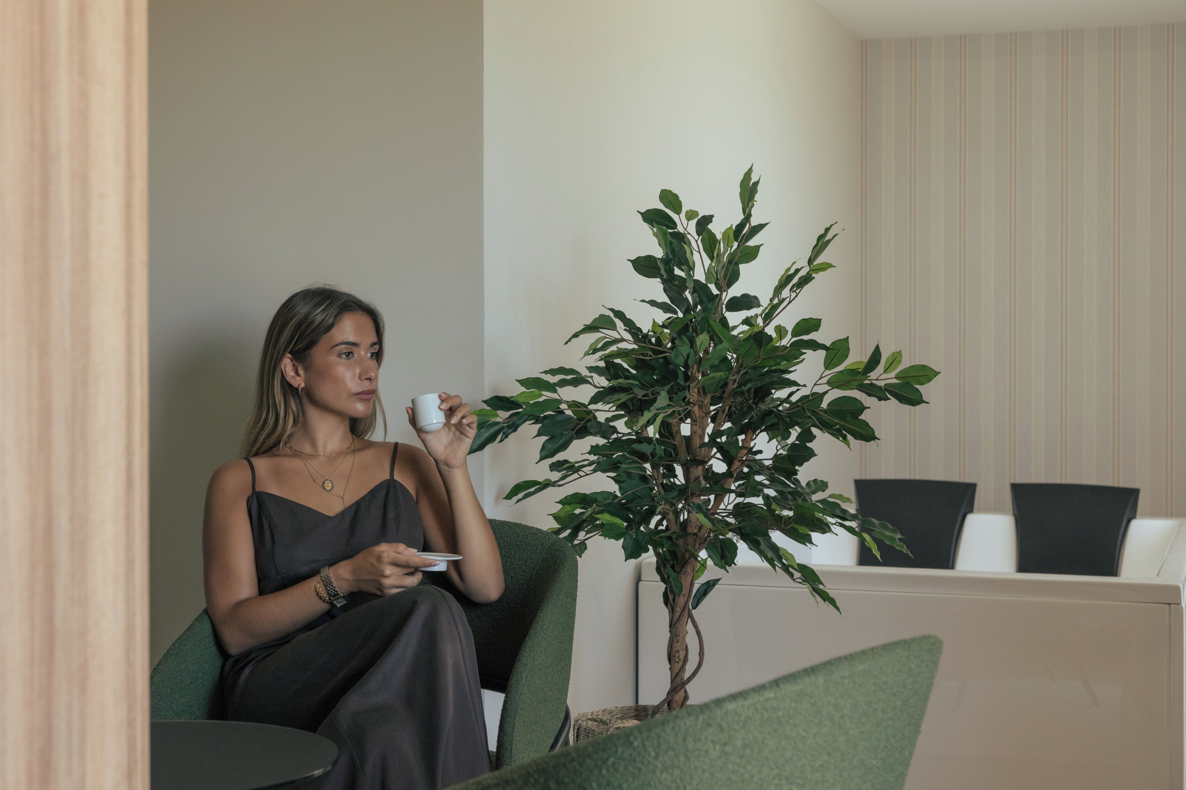 A woman seated on a green chair in a room with a potted plant, a white table, and a striped wall.