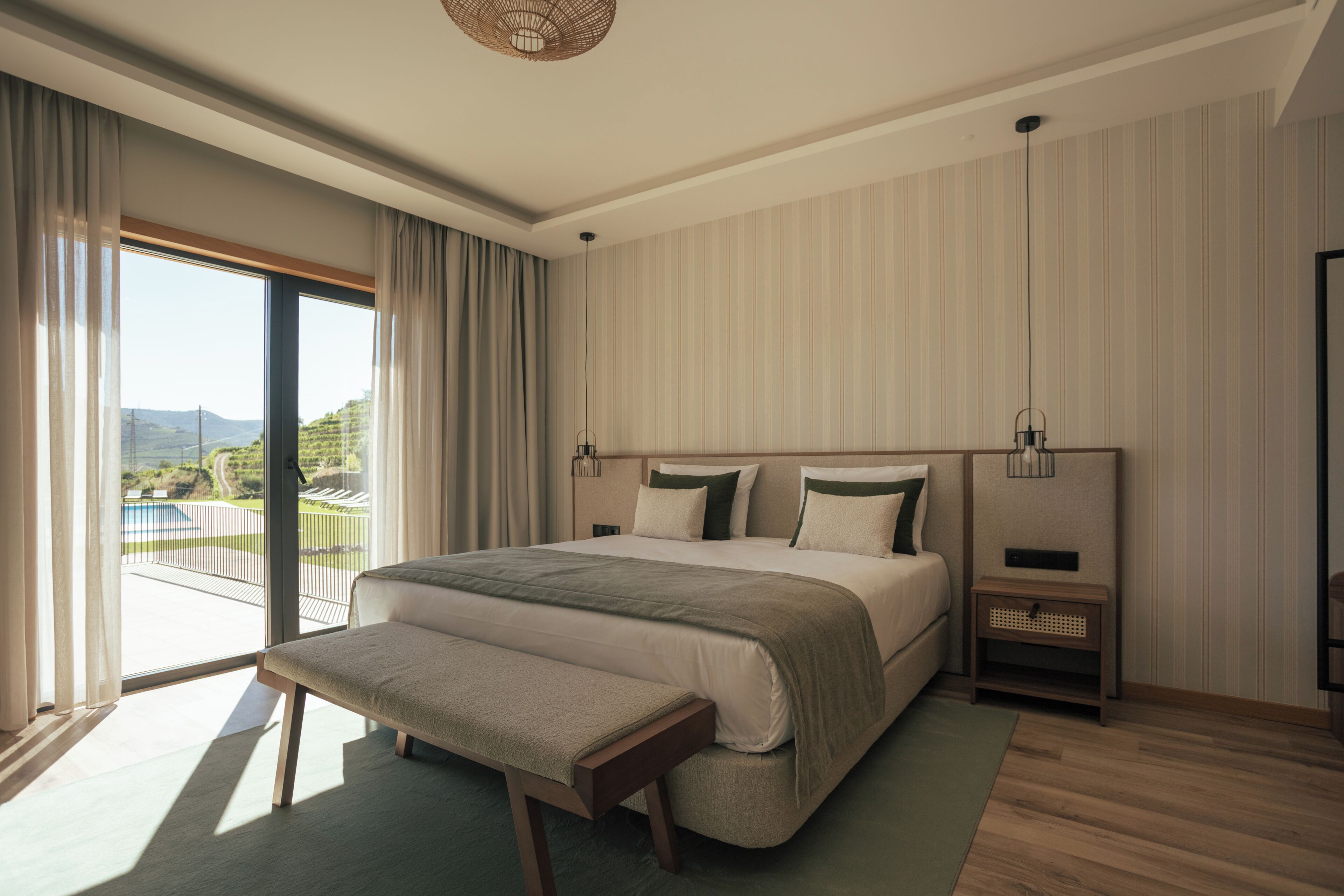 A modern bedroom with a large bed, a bench, a nightstand, and a view of a pool and mountains.