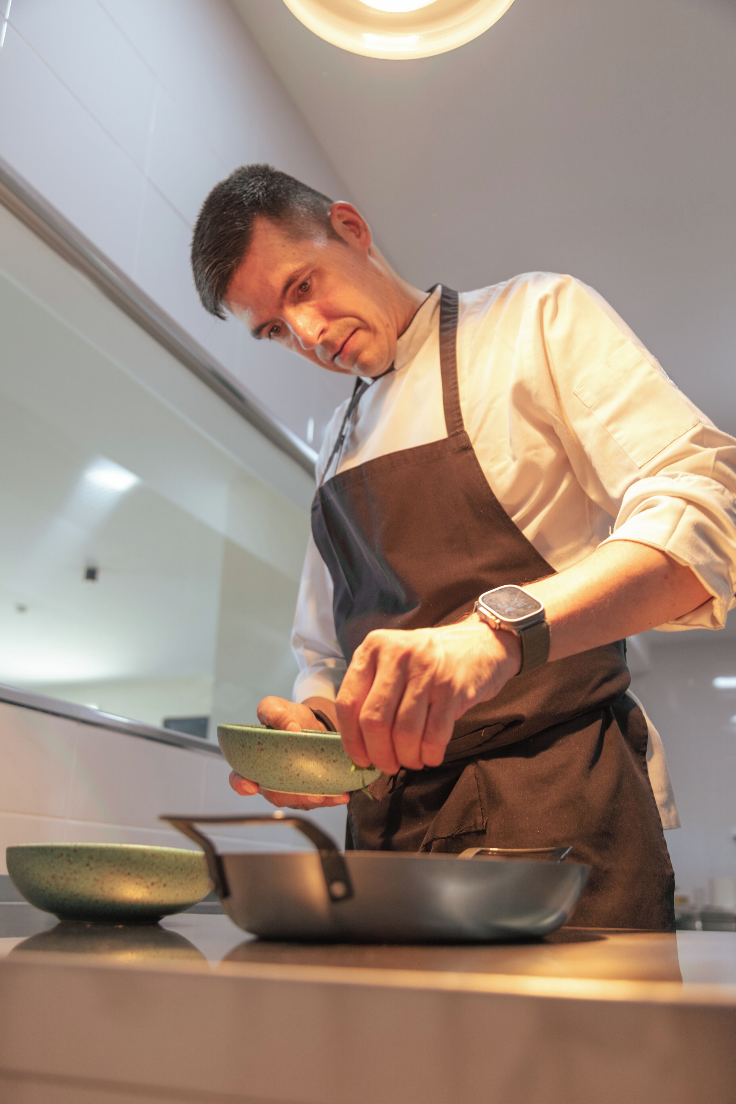 Uma pessoa em uma cozinha, vestindo uma camisa branca e avental marrom, segurando uma tigela verde sobre uma panela.
