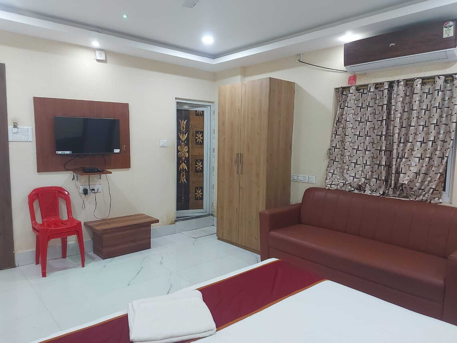 Quarto com televisão na parede, uma cadeira de plástico vermelha, um armário de madeira, um sofá castanho e uma cortina estampada.