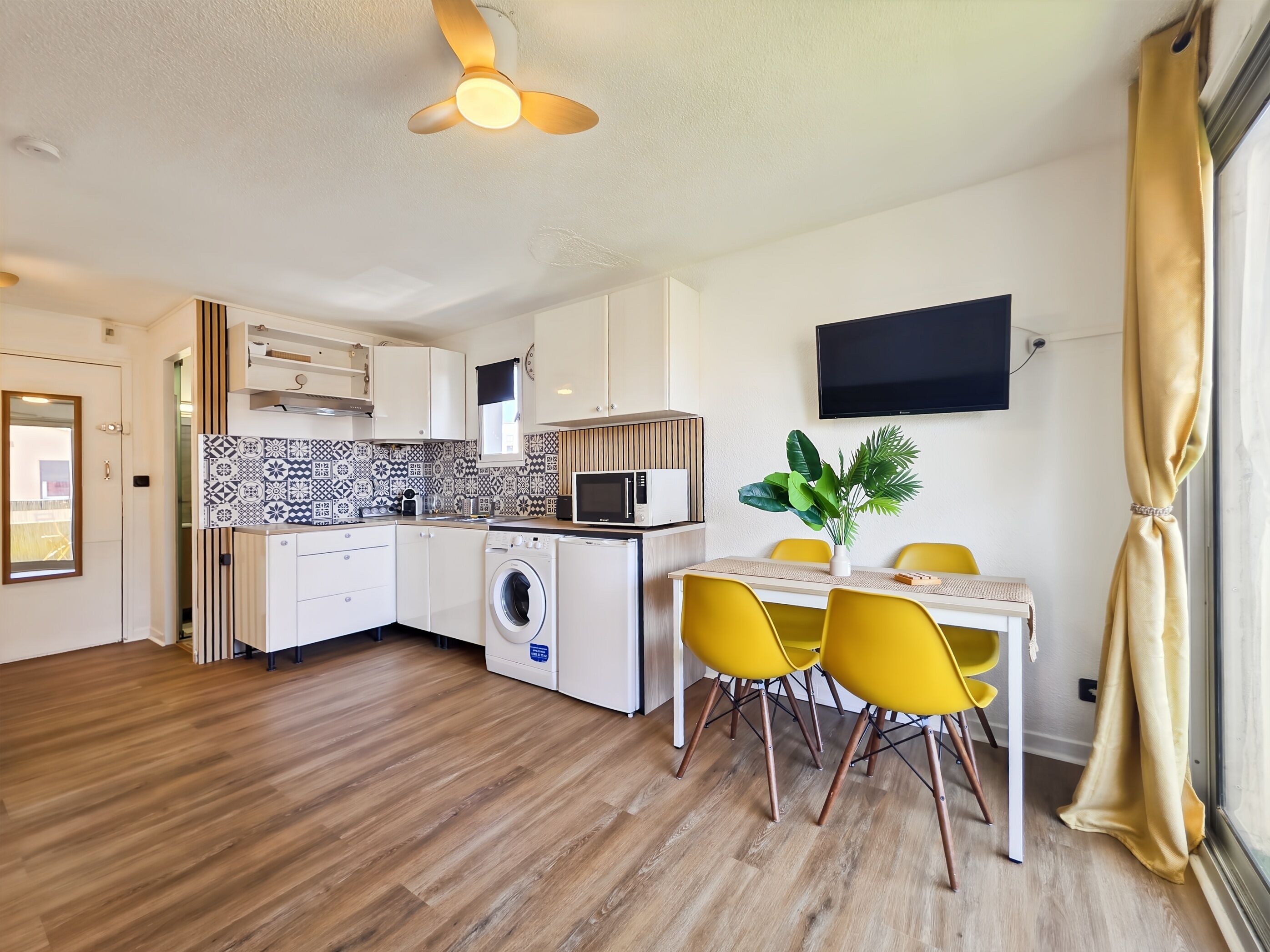 A modern kitchen with a washing machine, a dining area with yellow chairs, and a ceiling fan.