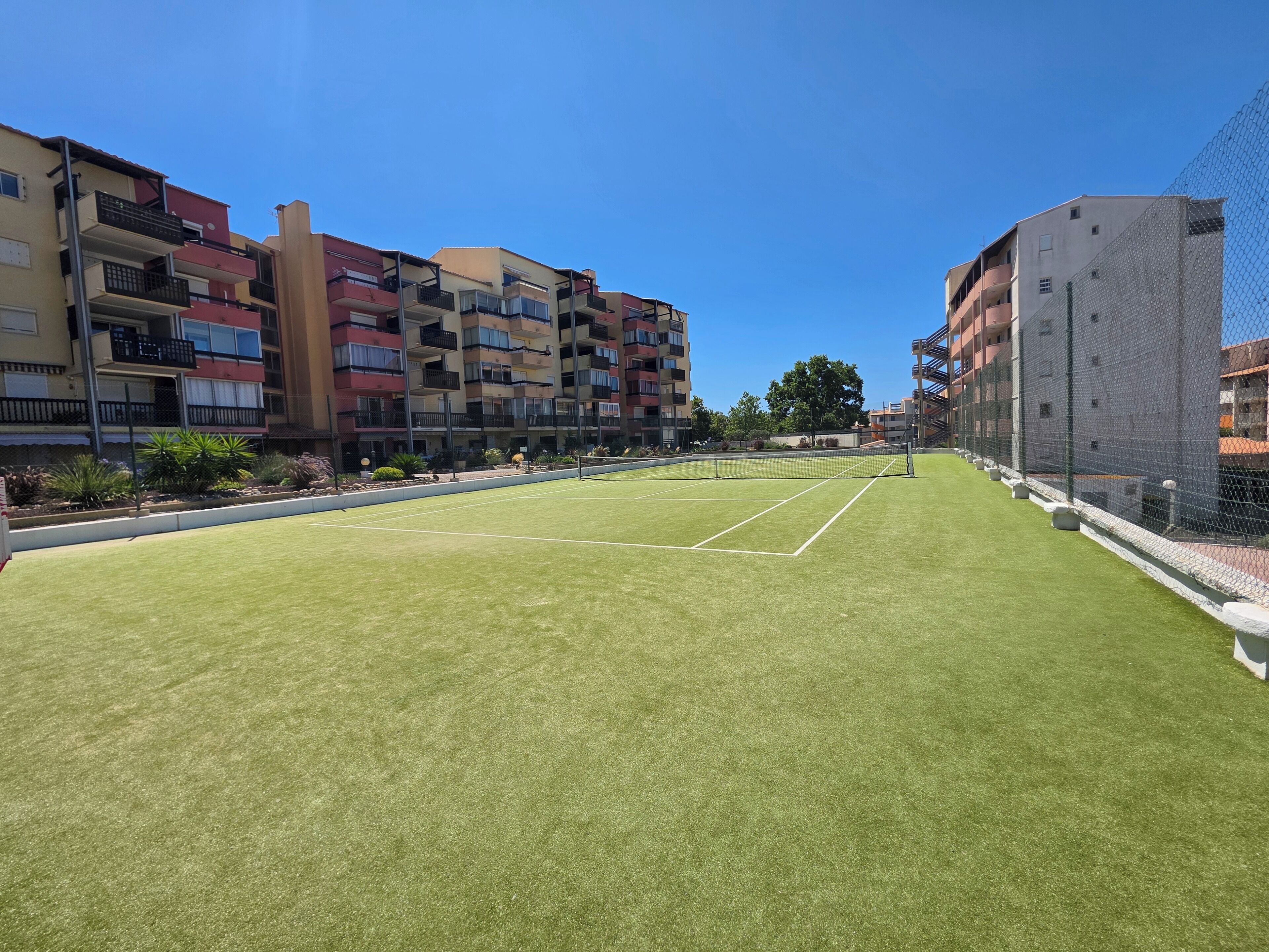 A tennis court with a net, surrounded by a chain-link fence, and adjacent multi-story buildings.