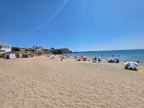 Platja sorrenca amb ombrel·les, algunes persones i un edifici amb teulada al fons.