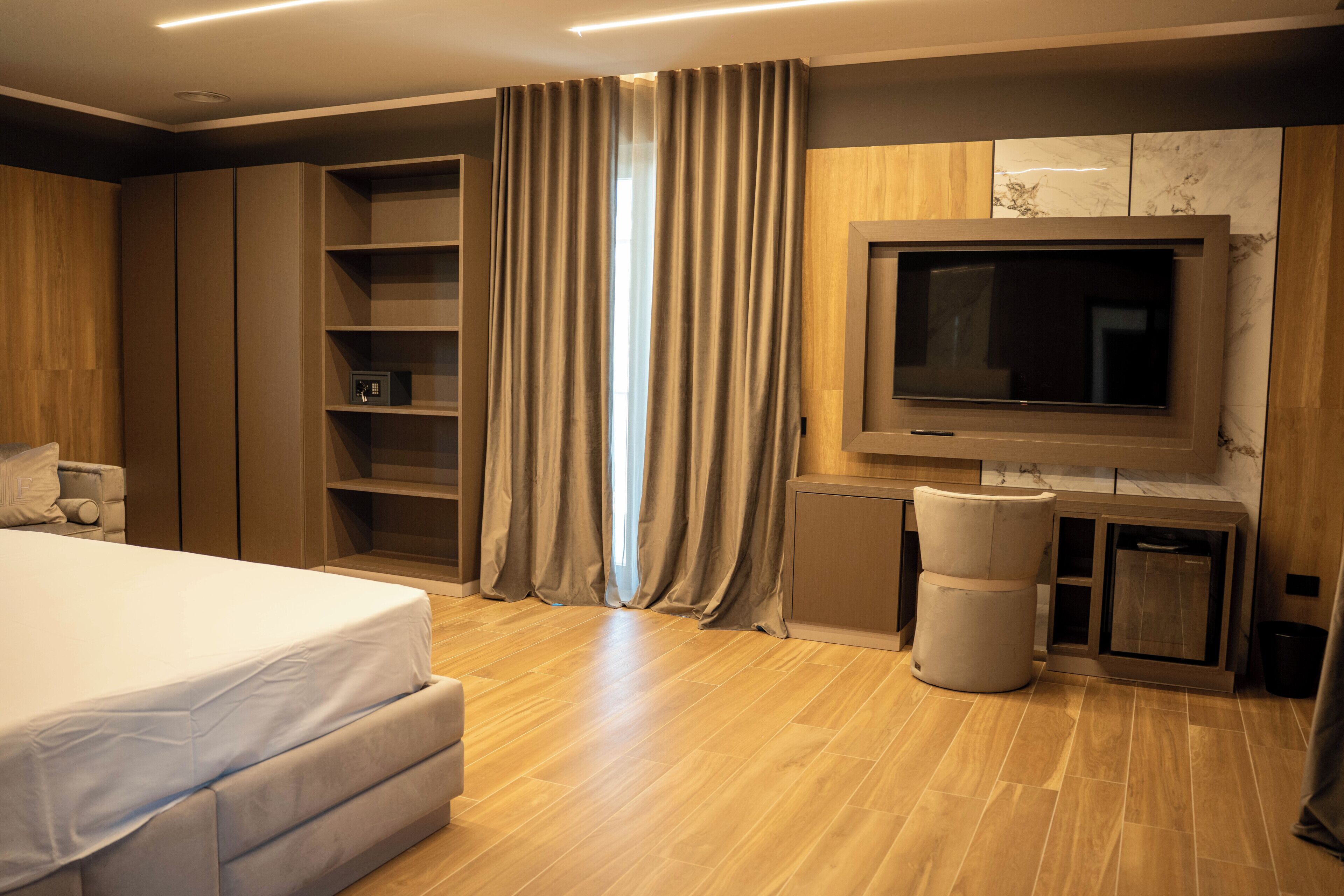 A modern bedroom with a large bed, built-in wardrobe, TV, and a small seating area.