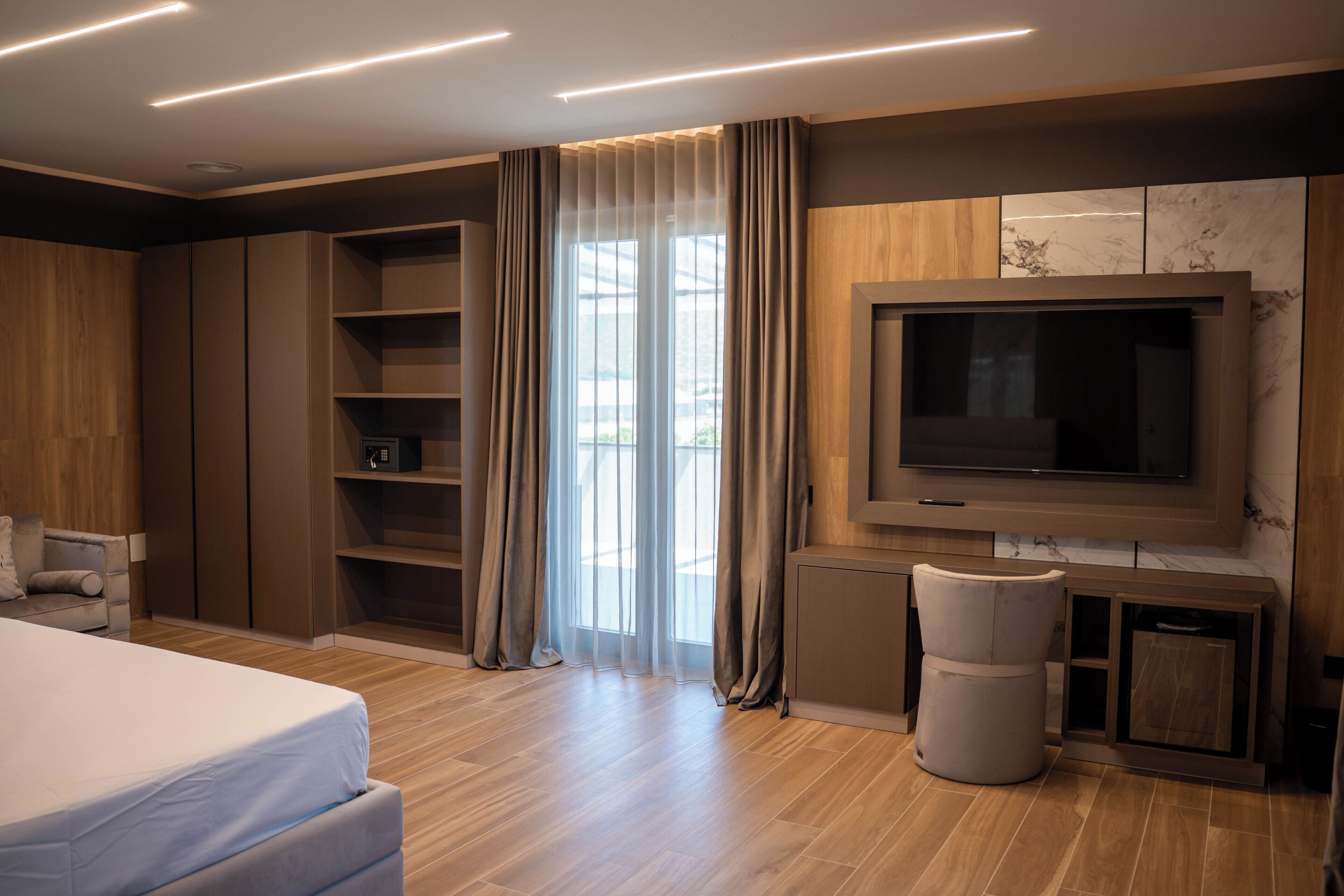 A modern bedroom with a bed, built-in shelving, a TV, and a large window with curtains.