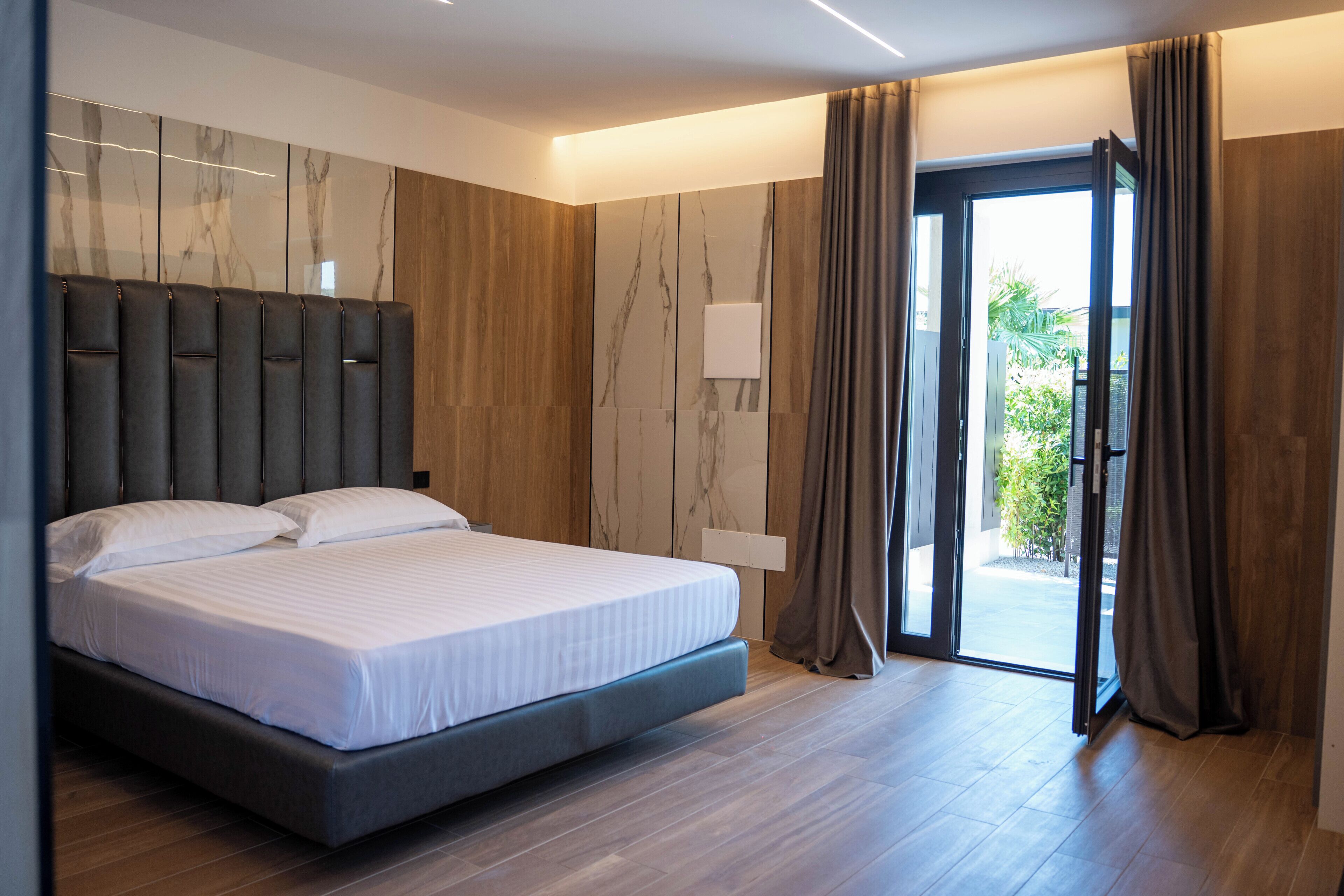 A modern bedroom with a large bed, a wall-mounted headboard, a sliding glass door to the outside, and a wooden floor.