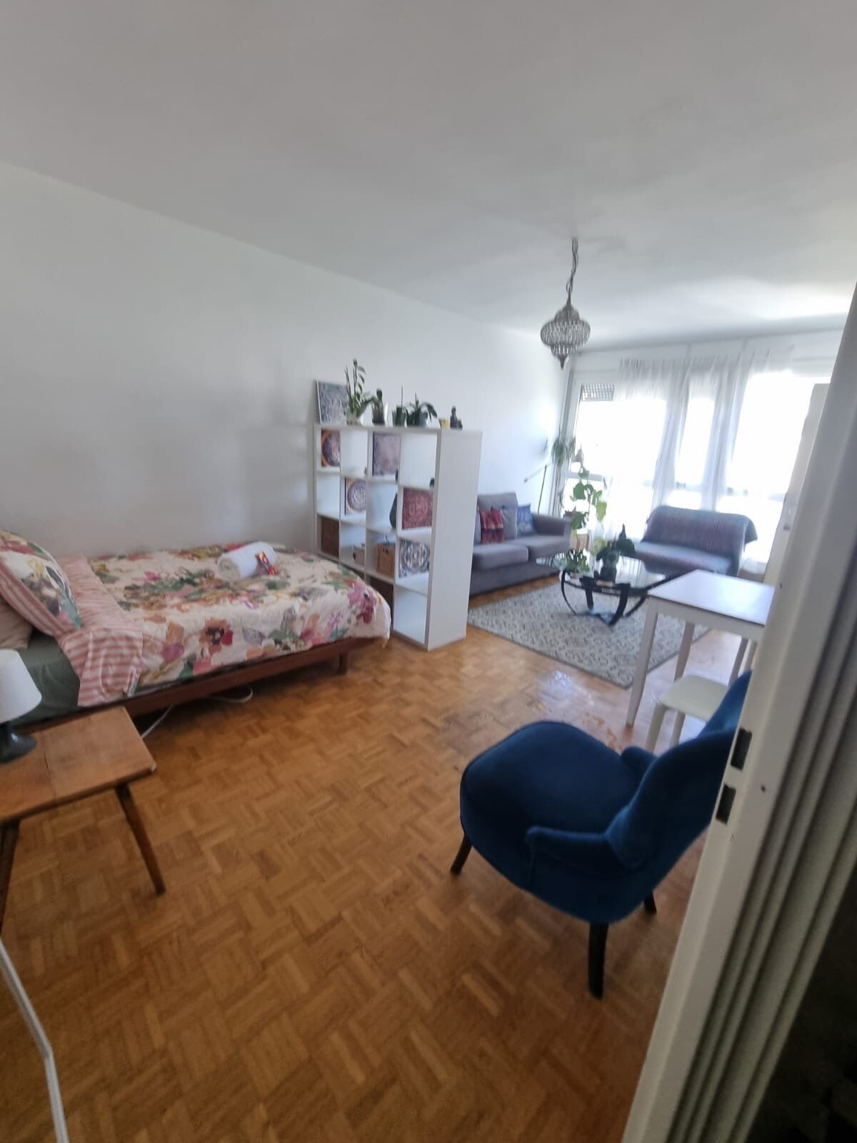 A living room with a bed, a blue chair, a white bookshelf, and a dining area.
