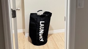 Quarto com piso de madeira, porta branca, parede branca e uma sacola de roupa suja preta com a inscrição 'LAUNDRY'.