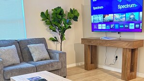 Sala de estar com televisão de tela plana exibindo a interface de um serviço de streaming, um sofá cinza, uma mesa de centro de madeira e um vaso com planta.