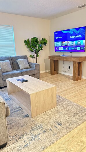 Ein Wohnzimmer mit Flachbildfernseher, der eine Streaming-Dienstanwendung zeigt, einem grauen Sofa, einem Holztisch und einer Topfpflanze.