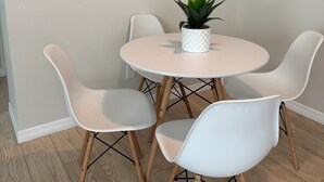 Uma sala de jantar com uma mesa redonda, quatro cadeiras brancas, um vaso de planta e um relógio fixado na parede.