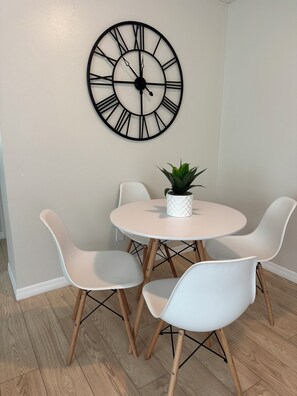 Ein Essbereich mit einem runden Tisch, vier weißen Stühlen, einer Topfpflanze und einer an der Wand befestigten Uhr.