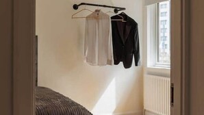 Un dormitorio con una cama, una ventana, un radiador y un perchero con una chaqueta y una camisa.