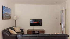 Una sala de estar con un sofá de cuero negro, un televisor montado en la pared y un cuadro con una imagen de un estadio.